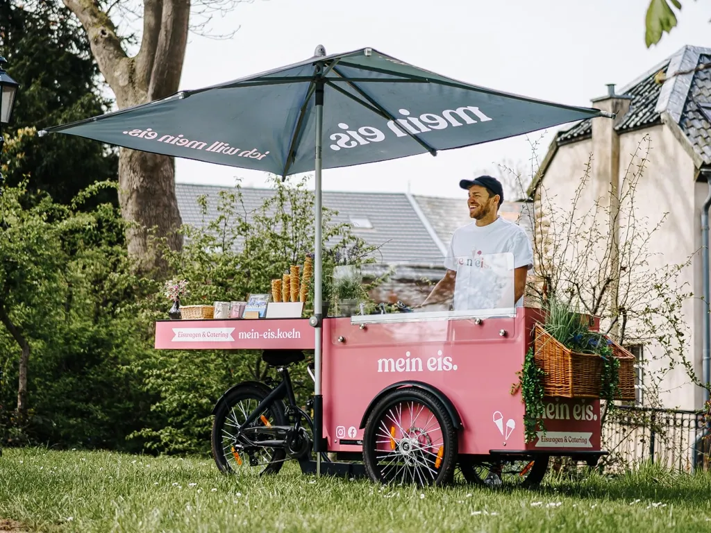 Rosa Eisfahrrad von mein eis mit Schirm und Servicemitarbeiter auf einer Wiese vor einem Haus